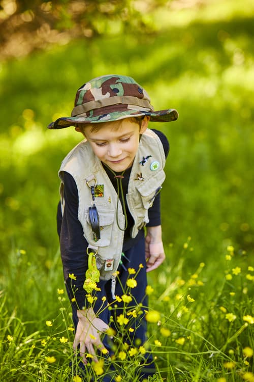 A young boy in a hunting vest and cap admires yellow mustard flowers